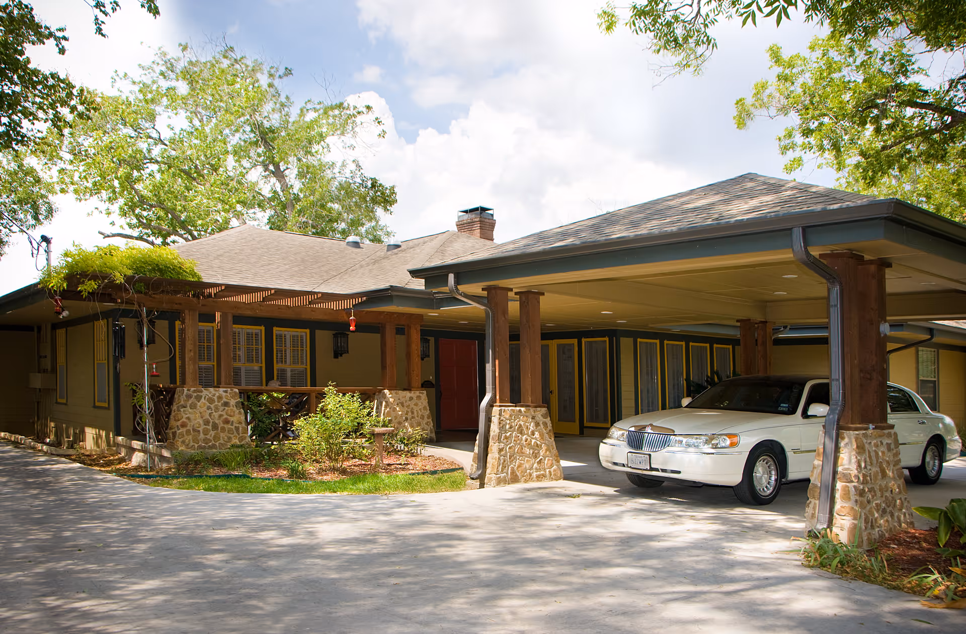 Exterior view of a single-story assisted living facility with a covered driveway supported by stone and wooden pillars. Two white cars are parked under the covered area. The building has a porch with stone accents and greenery around it, with trees and a partly cloudy sky in the background.