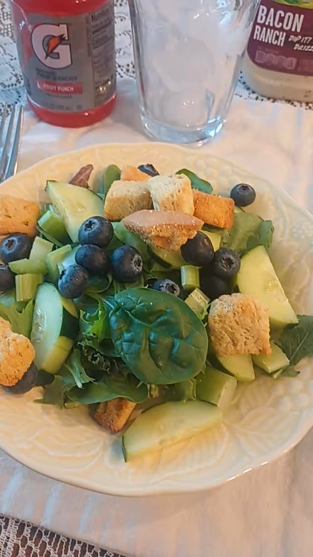 Plate of mixed greens salad with cucumbers, blueberries, and croutons atop a table