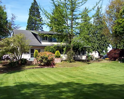 Two-story residence with a large manicured lawn, trees, and shrubs in front under a blue sky.