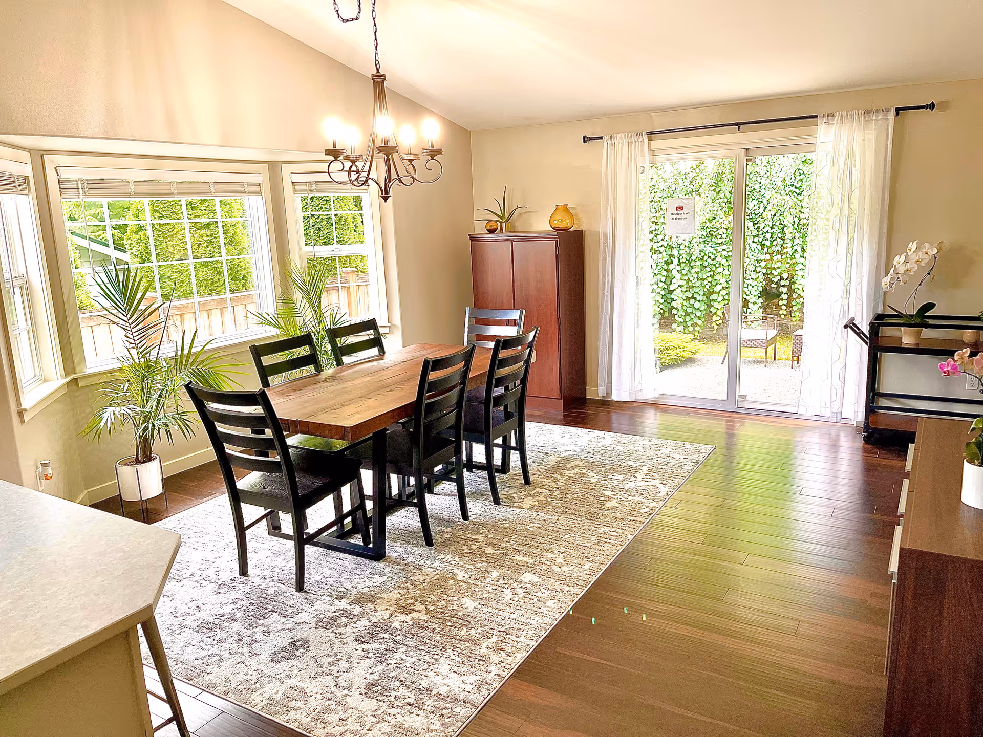 Bright dining room with a wooden table and chairs, chandelier, plants, and sliding glass door.