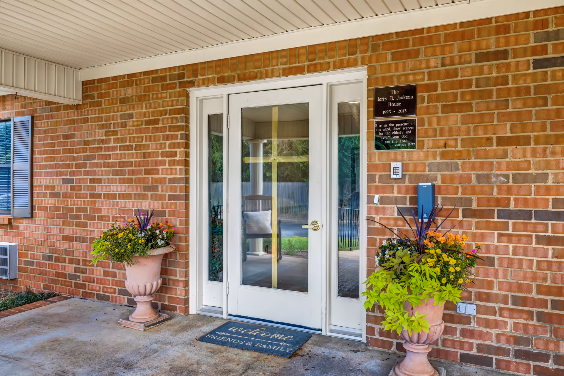 Brick building entrance with white double glass doors flanked by potted plants and a welcome mat.