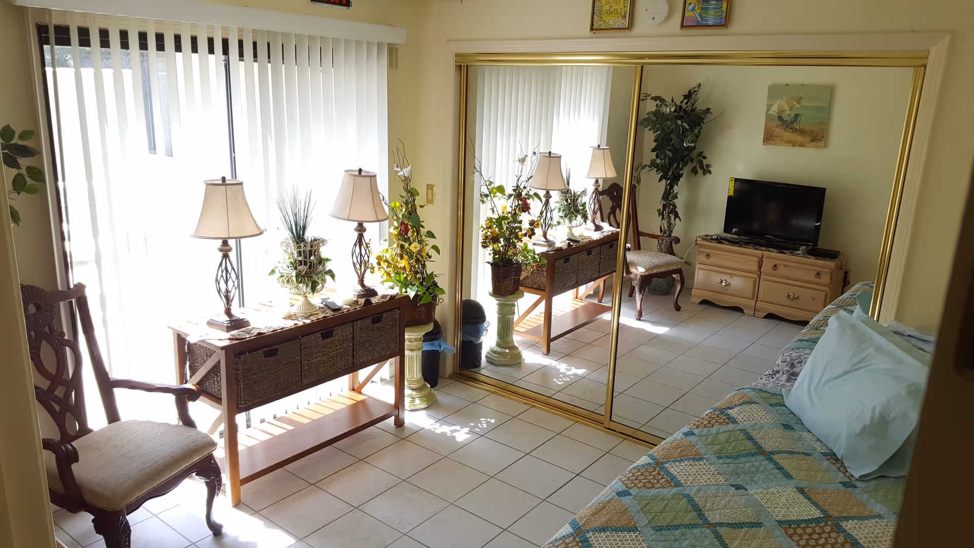 Sunlit bedroom with bed, mirrored closet doors, dresser, and seating area in a senior facility.