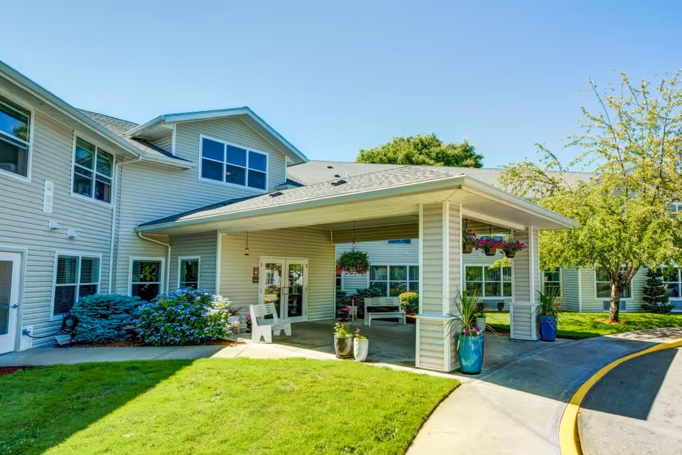 Front entrance of an assisted living facility with a covered drop-off portico, potted plants, benches, and a manicured lawn.