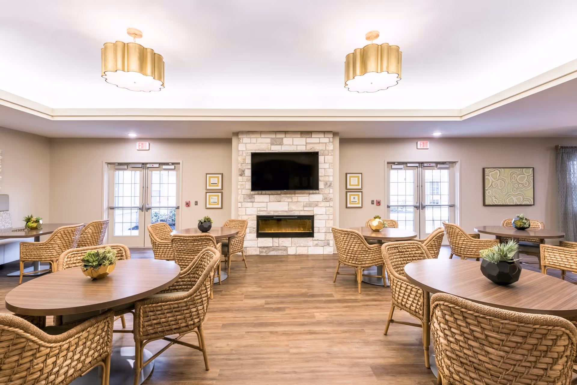 A bright and spacious common area with multiple round wooden tables surrounded by wicker chairs. Each table has a small decorative plant. The room features a stone fireplace with a mounted flat-screen TV above it. There are two sets of double glass doors on either side of the fireplace, and the ceiling has recessed lighting with two large, decorative pendant lights. The floor is wood, and the walls are painted a soft beige color with some framed artwork.