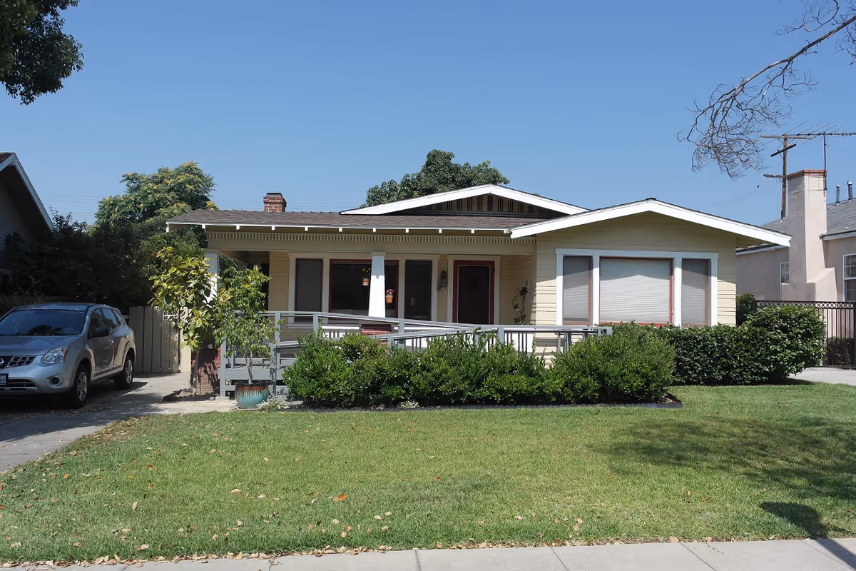 Single-story yellow house with a front porch and wheelchair ramp, shrubs and a parked car on a sunny day.