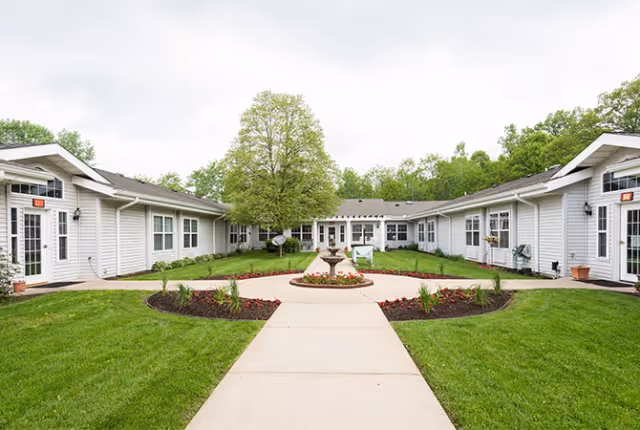 Courtyard of a single-story senior living facility with a central walkway, fountain, lawns, and surrounding white buildings.