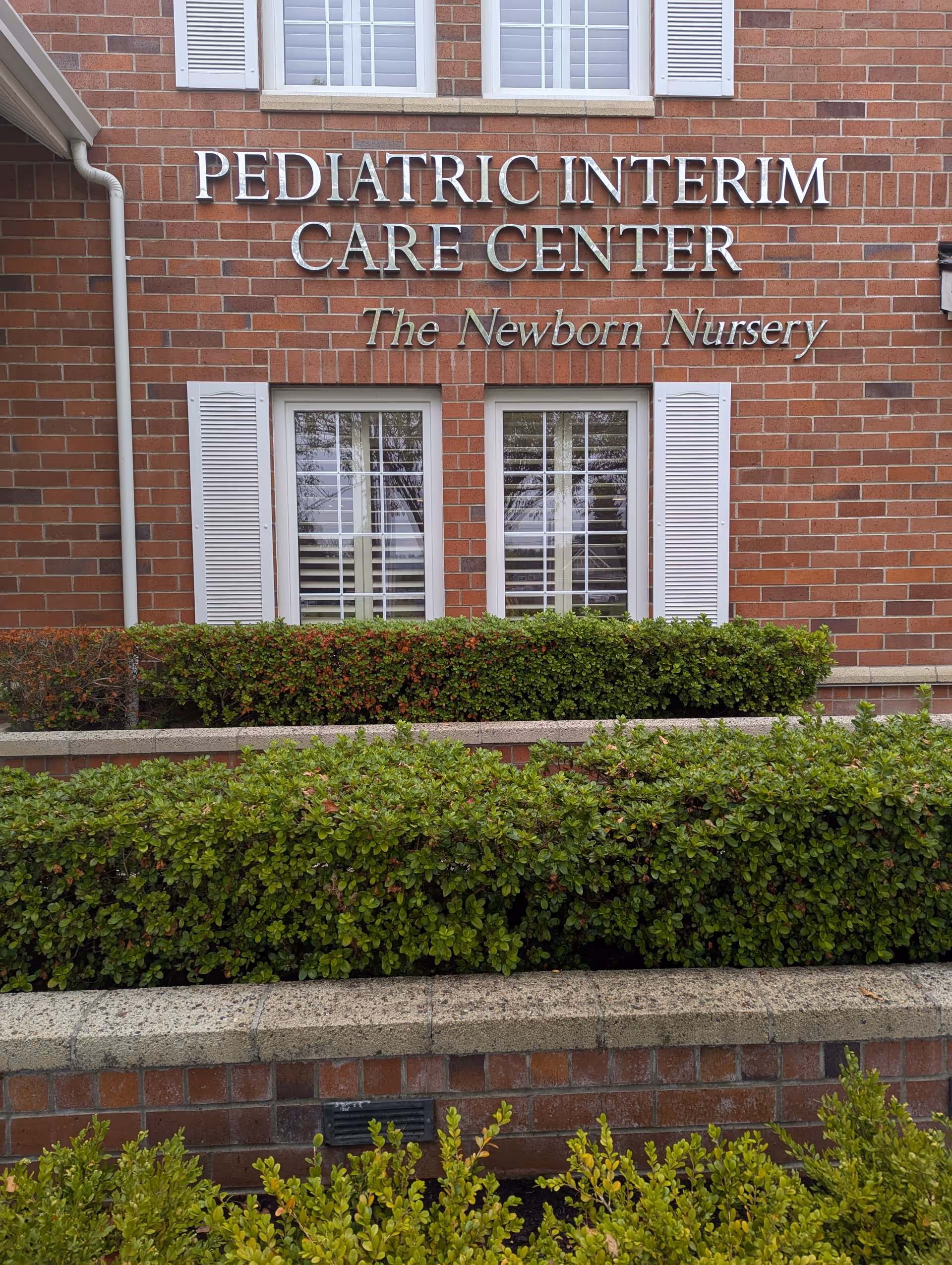 Red brick building exterior with 'Pediatric Interim Care Center' sign and shrubs.