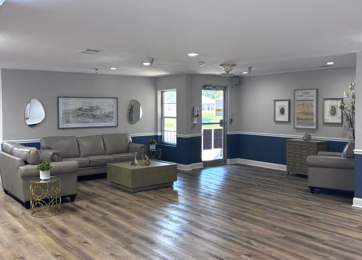 A spacious and well-lit living room area with light gray walls and a dark blue wainscoting. The room features two gray leather sofas, a large square coffee table, two small round side tables with plants, and framed artwork and mirrors on the walls. The floor is wood with a natural finish, and there is a glass door leading outside.