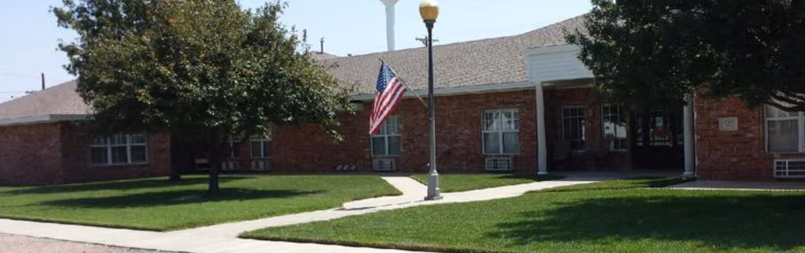 Exterior view of a single-story brick building with multiple windows, a sidewalk leading to the entrance, green grass, trees, a streetlamp, and an American flag on a pole.