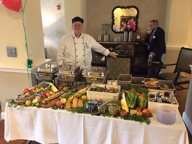 Chef in uniform oversees a buffet table with chafing dishes and fresh produce in dining area