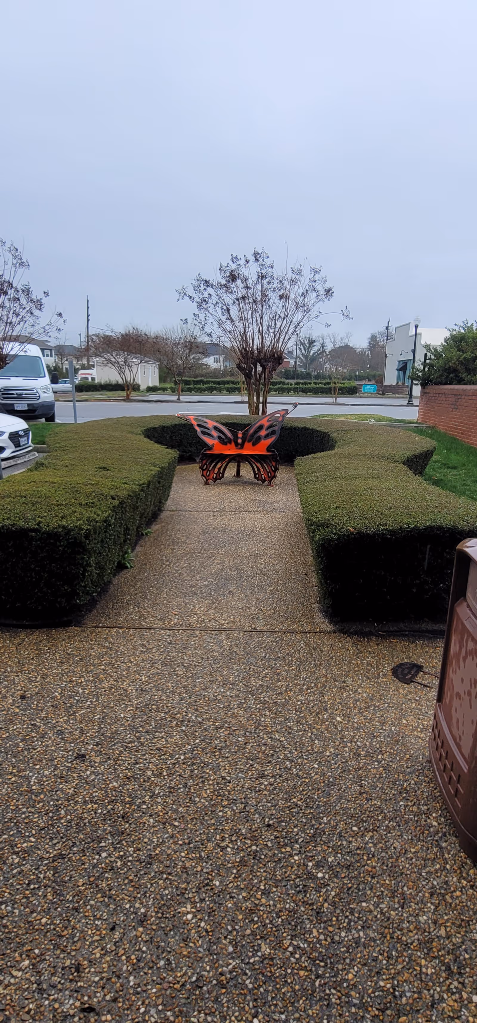Path bordered by neatly trimmed hedges leads to a butterfly sculpture in a landscaped outdoor area.
