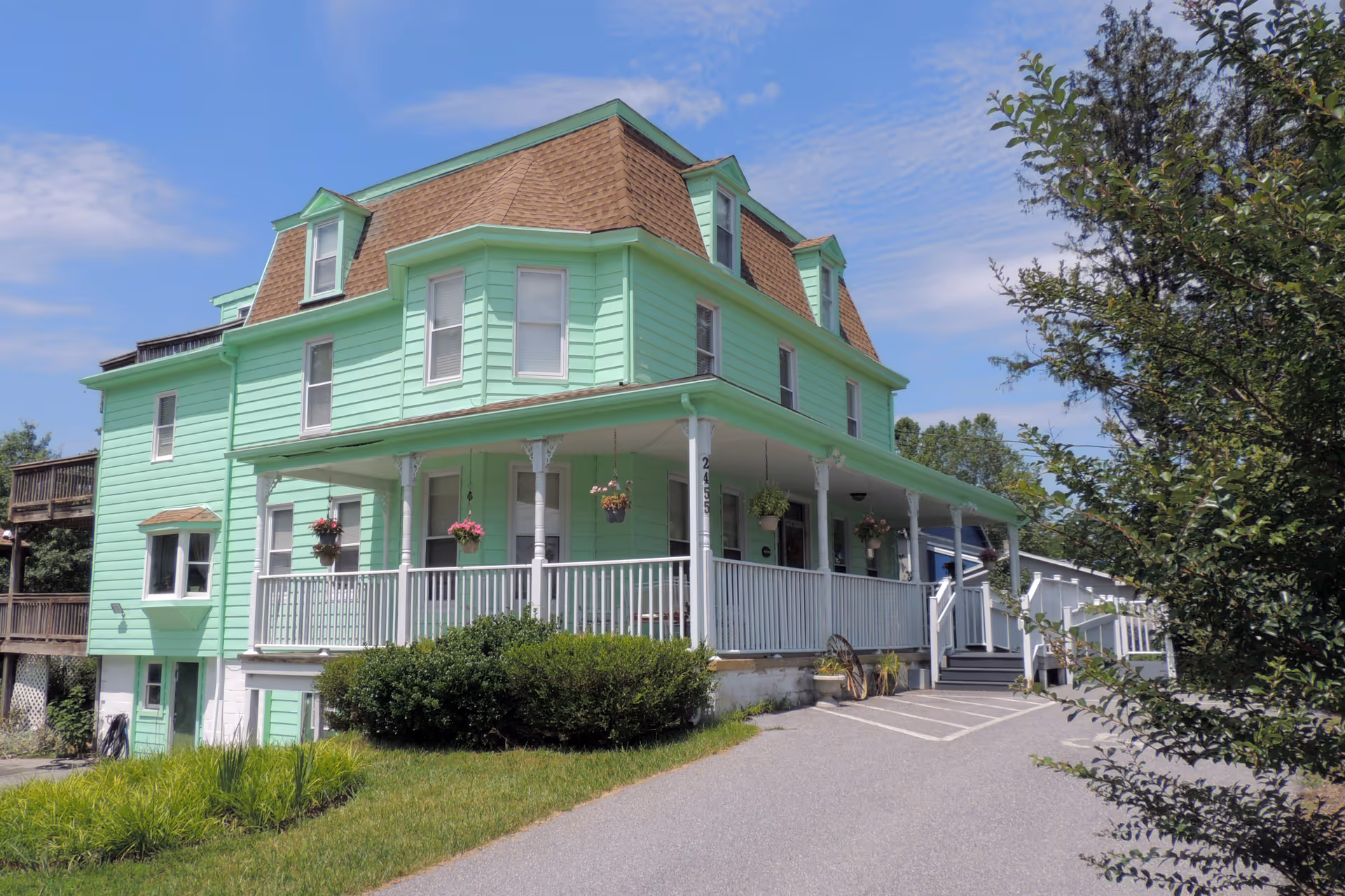 A large, three-story mint green house with a brown mansard roof and white porch railing. The house has several windows and hanging flower pots on the porch. There is a driveway leading up to the porch with greenery and trees surrounding the property under a partly cloudy blue sky.