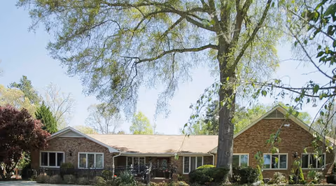 One-story brick senior living facility facade with a large tree and shrubs.