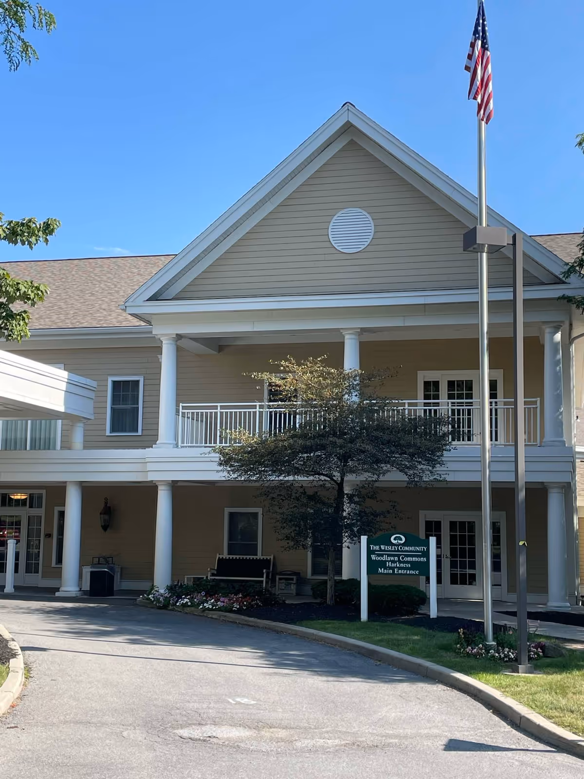 Front entrance of The Wesley Community — Woodlawn Commons building with a covered portico, white columns, flagpole, and a small green sign by the driveway.