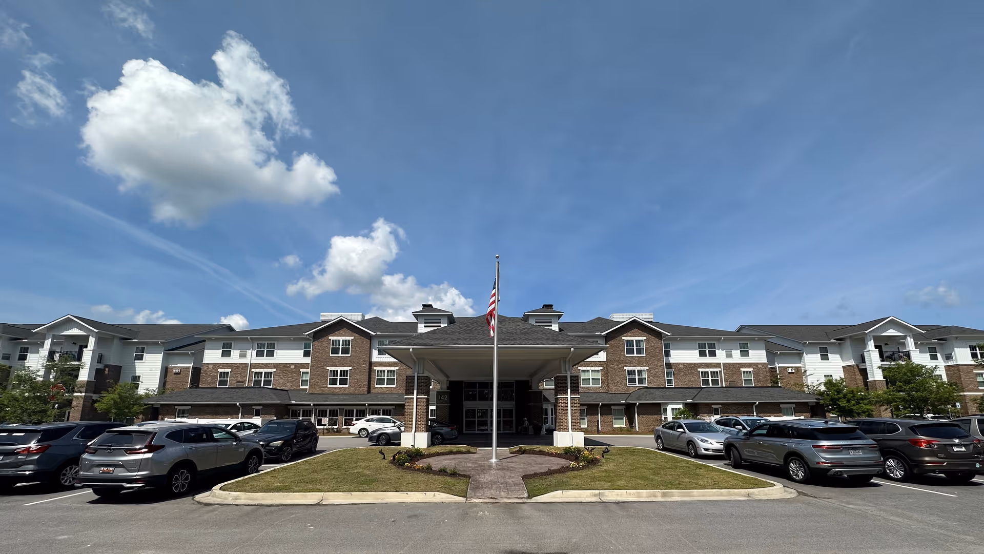 Front exterior view of Okatie Pines Retirement Community building with a covered entrance, an American flag on a flagpole in front, several parked cars, and a partly cloudy blue sky.