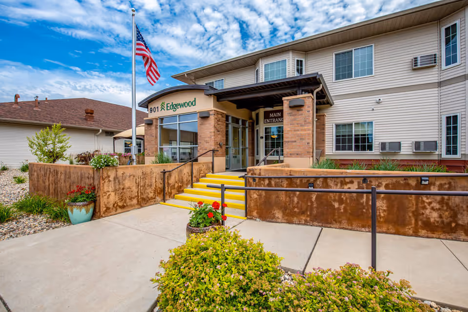 Front entrance of the Edgewood senior living facility showing the main entrance with a flagpole, yellow steps, wheelchair ramp, and landscaping.