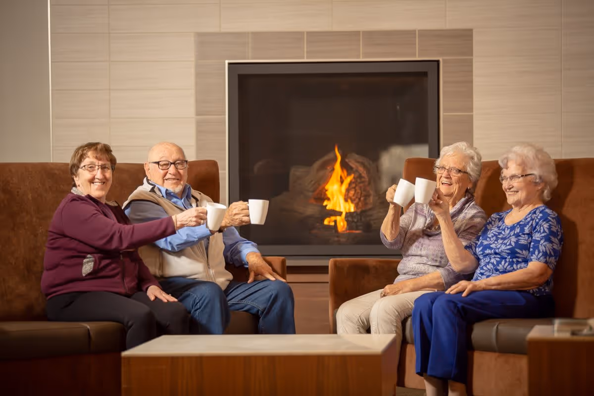 Four elderly people sitting on brown couches in front of a lit fireplace, smiling and holding white mugs as if toasting.