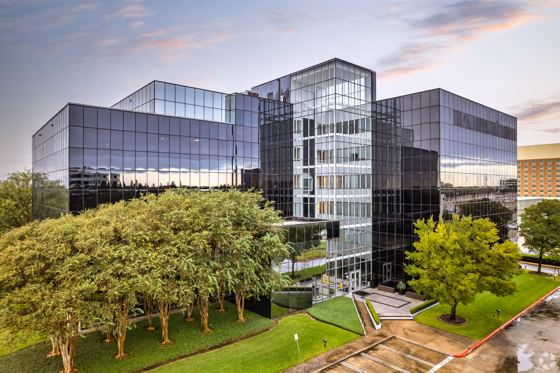 Glass-fronted modern building with reflective facade, trees, and a parking lot in front.