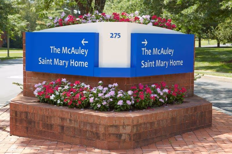 Brick planter with flowers and a blue directional sign reading 'The McAuley Saint Mary Home' by a tree-lined driveway.