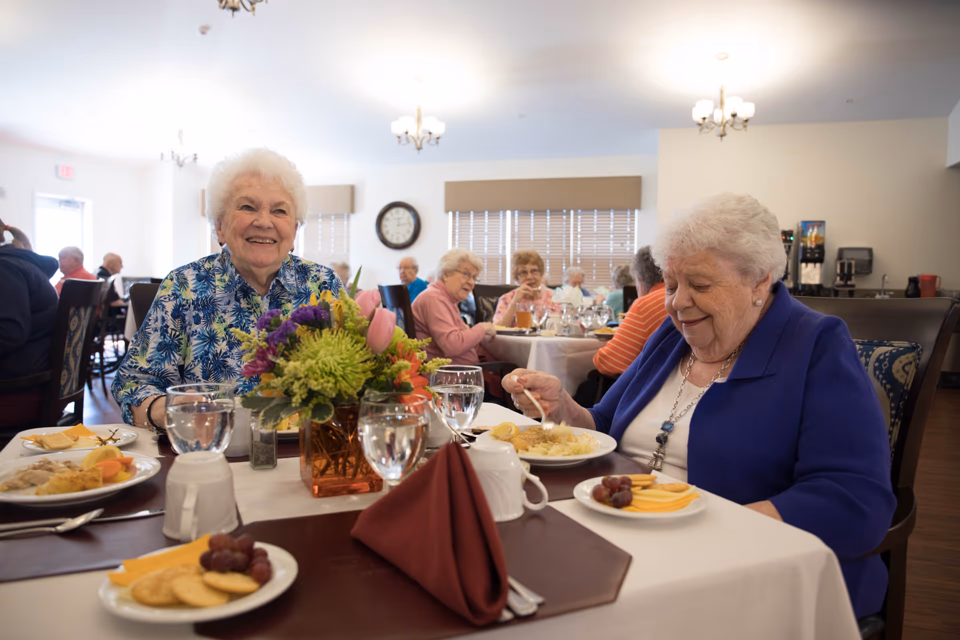 Two elderly women smiling and eating at a decorated dining table in a communal dining room with other residents in the background.