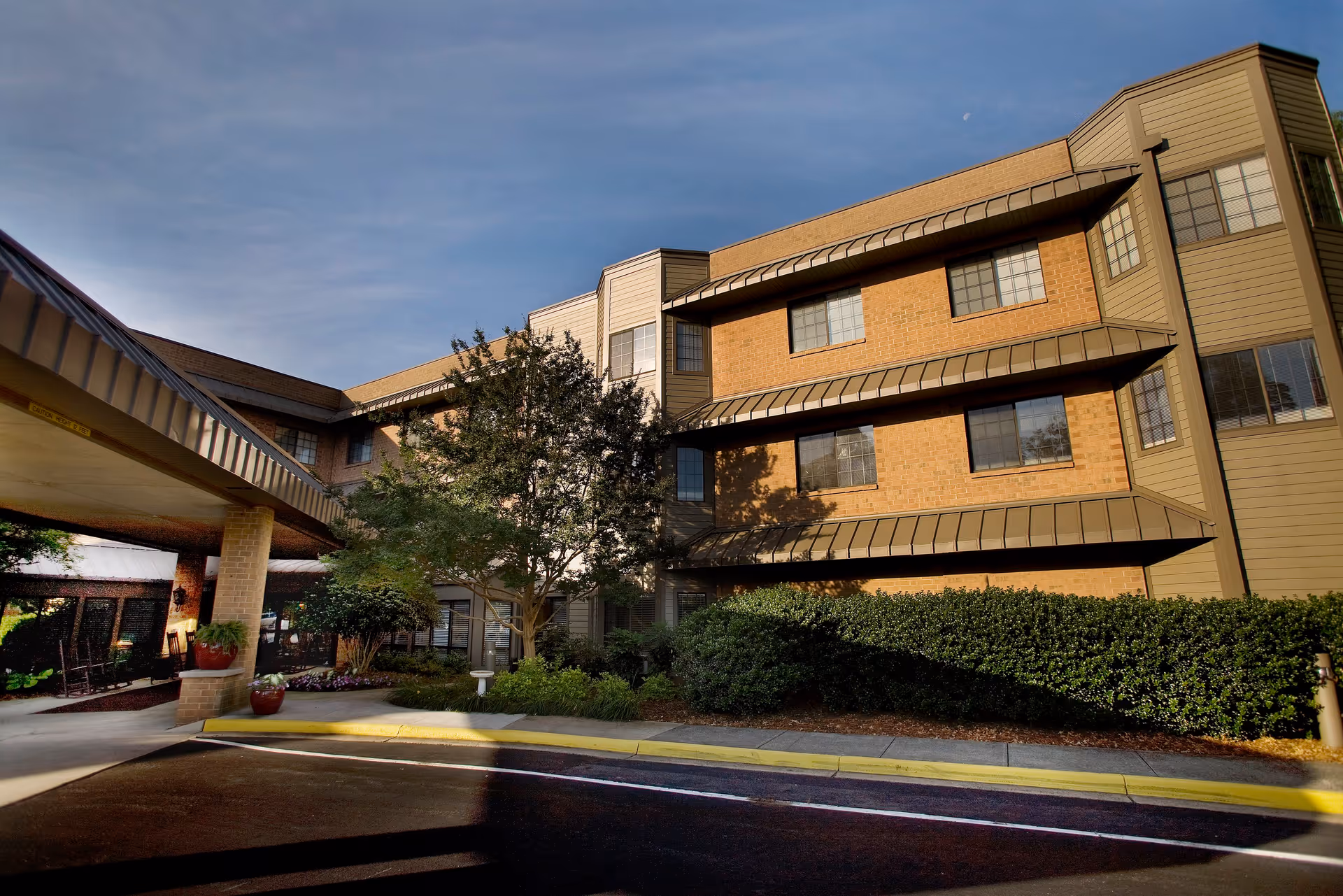 Exterior view of a multi-story senior living facility building with brick and siding walls, several windows, a covered entrance, and landscaped greenery including bushes and a tree.