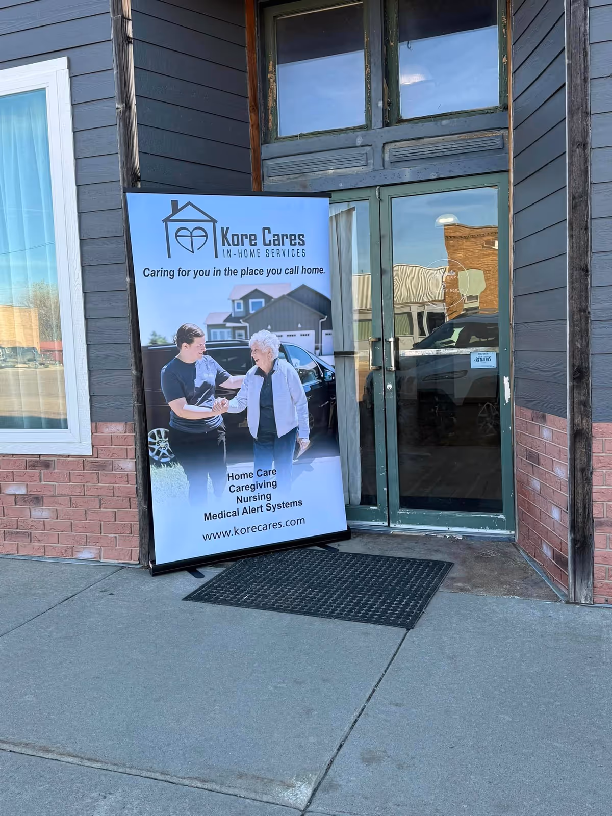 Exterior entrance with Kore Cares signboard showing a caregiver assisting an elderly person