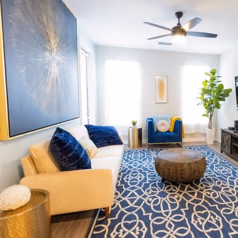 Bright modern living room with a cream sofa, blue armchair, patterned blue rug, round woven ottoman, large wall art, and ceiling fan.