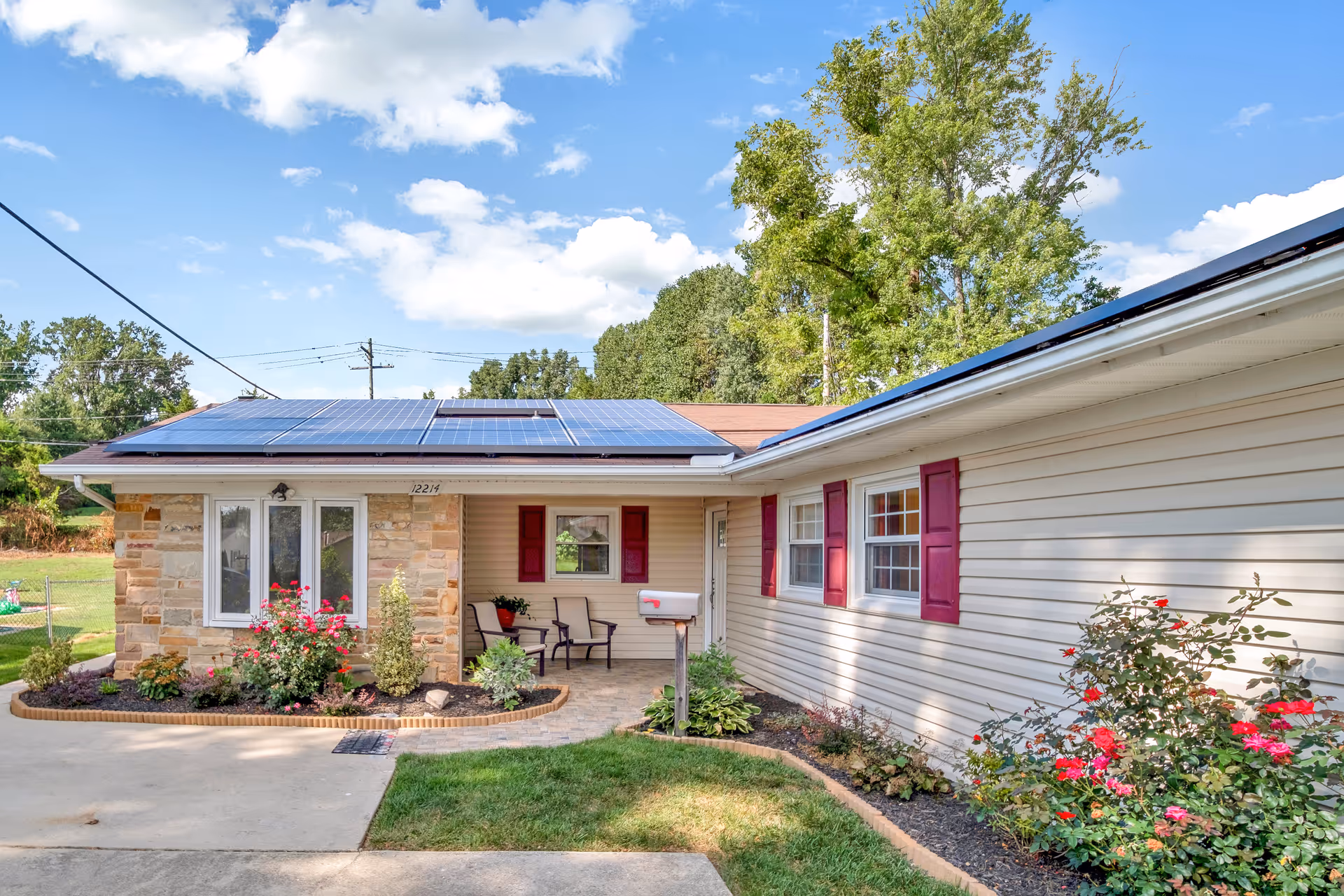 Single-story ranch-style building with solar panels on the roof, a small covered porch with chairs, red shutters, and landscaped flower beds.