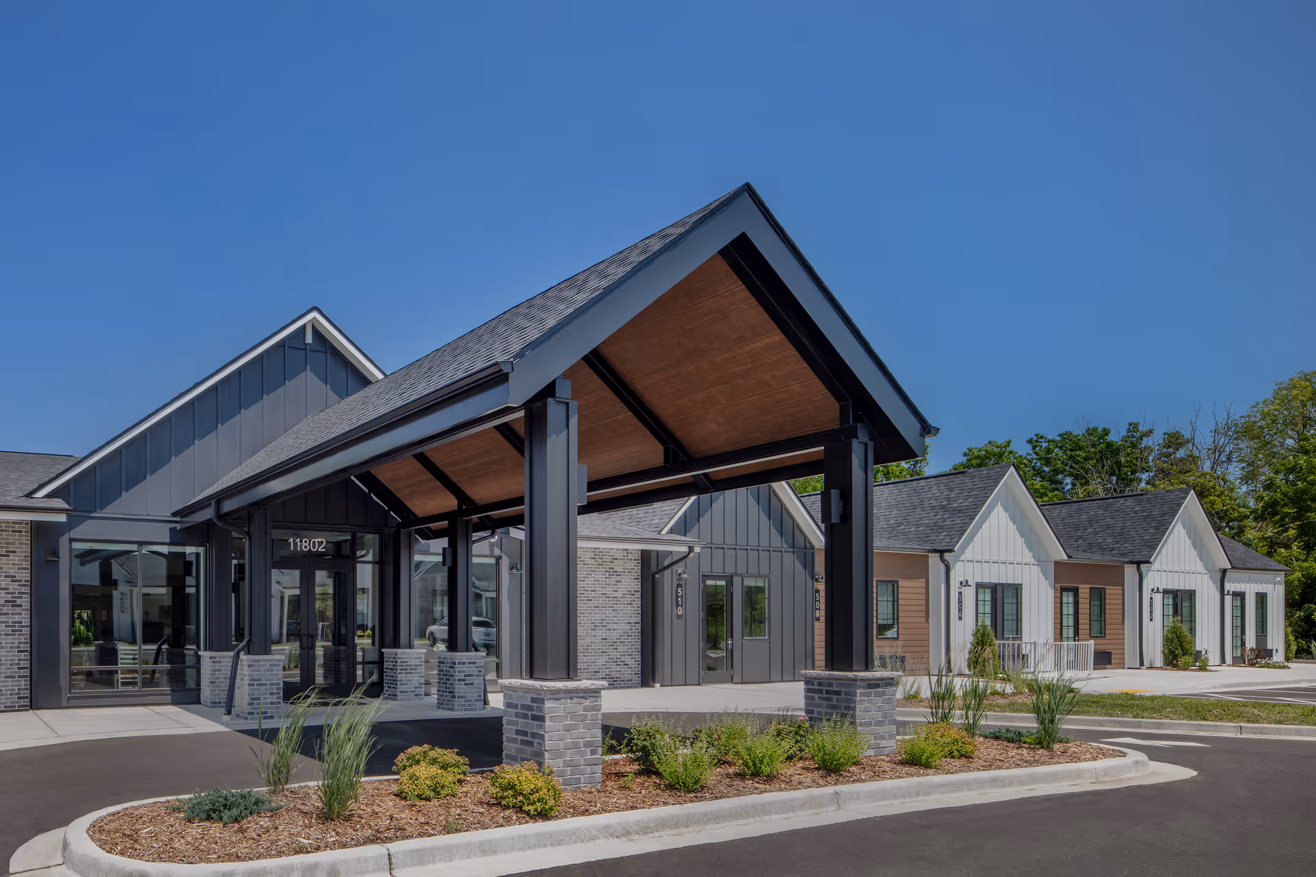 Exterior view of a modern senior living facility with a covered entrance supported by black pillars and brick bases. The building features a combination of gray and white siding with large windows and a neatly landscaped area with shrubs and plants in front. The sky is clear and blue.