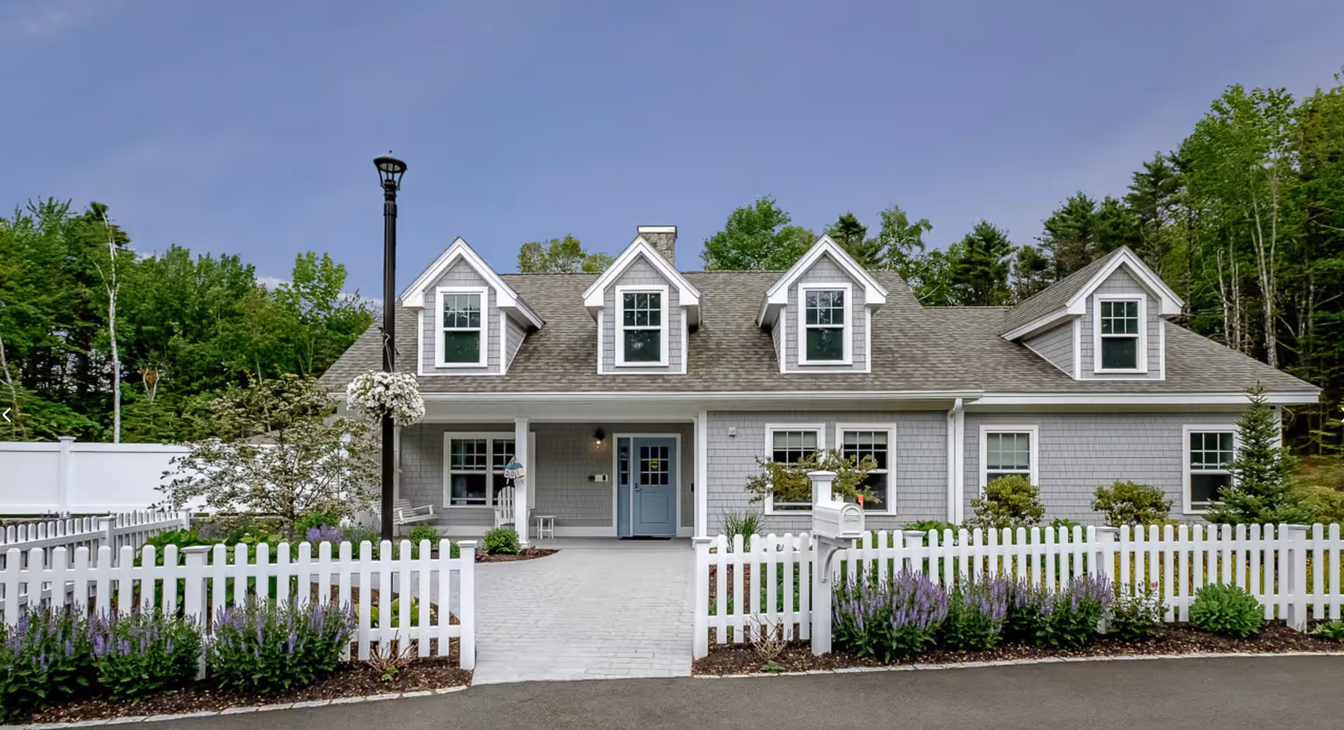 Front view of a gray two-story house with dormer windows, a white picket fence, mailbox, and landscaped yard.