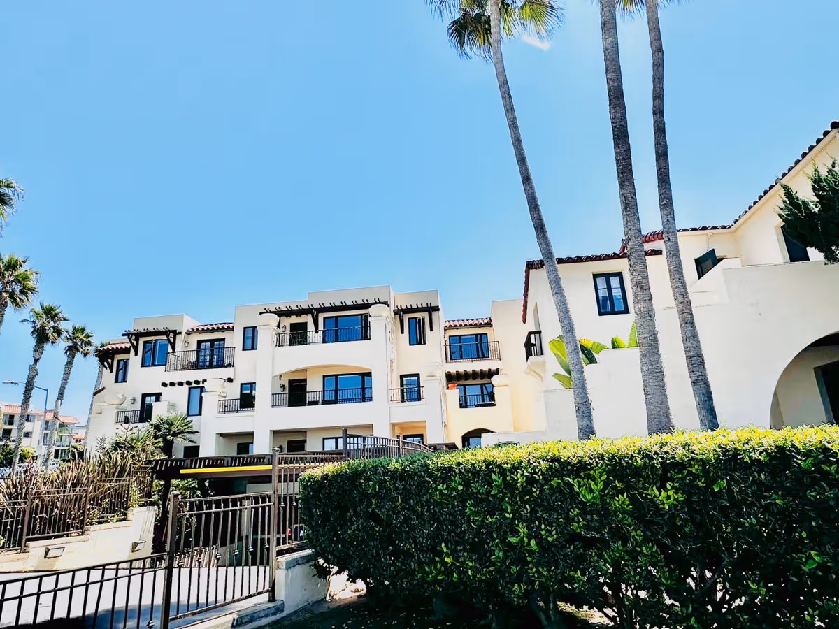 White Spanish-style multi-story building with balconies, palm trees, and a hedge in front under a clear blue sky.