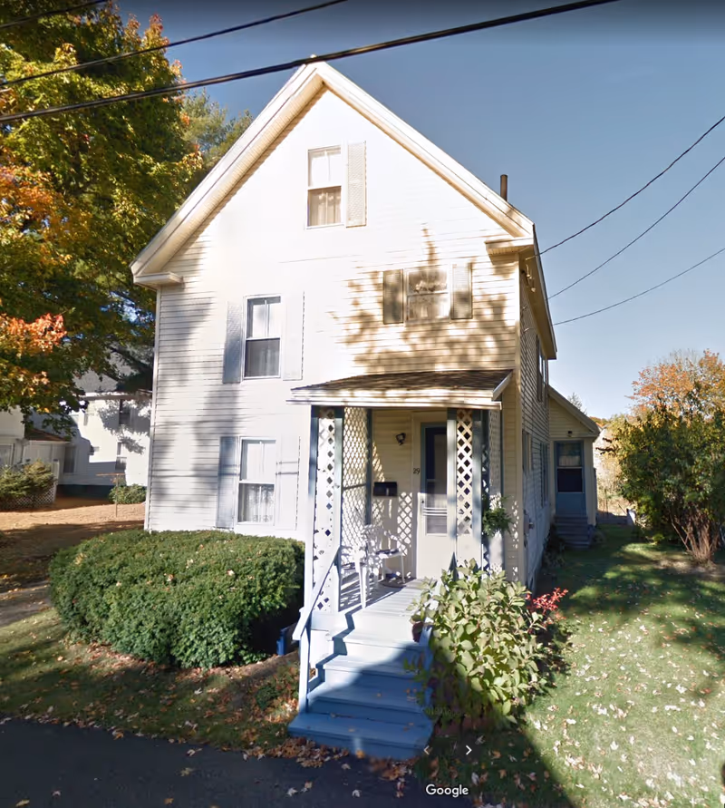 White two-story house with porch, steps, and surrounding shrubbery on a sunny day.