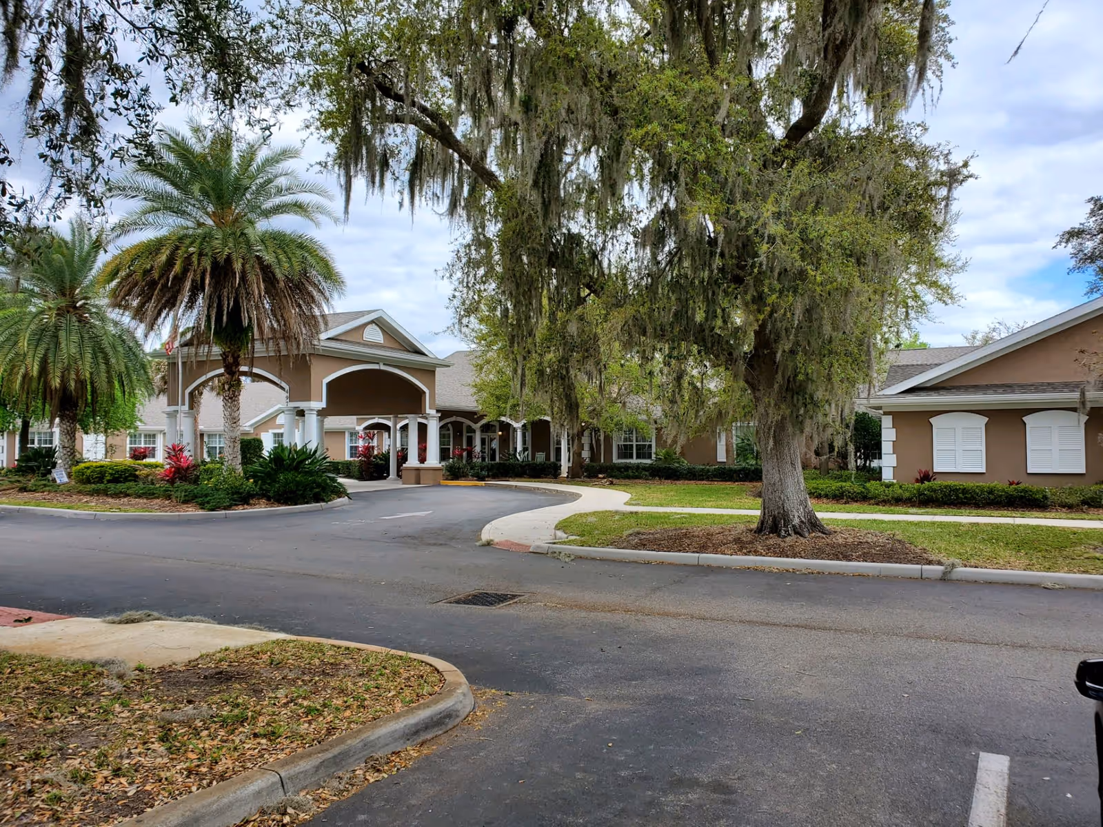 Exterior view of Addington Place of Titusville showing the entrance with a covered driveway, surrounded by palm trees, other greenery, and a large tree with hanging moss. The building is beige with white trim and has a well-maintained landscape.