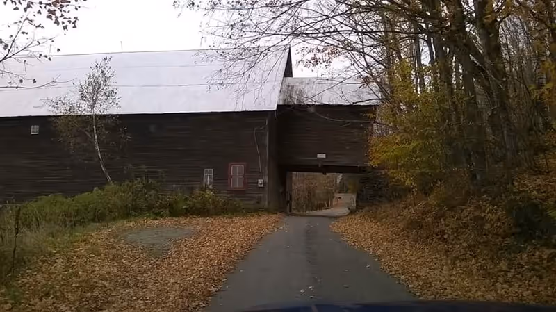 Rustic wooden building with a covered passageway, driveway, and autumn foliage along the roadside.