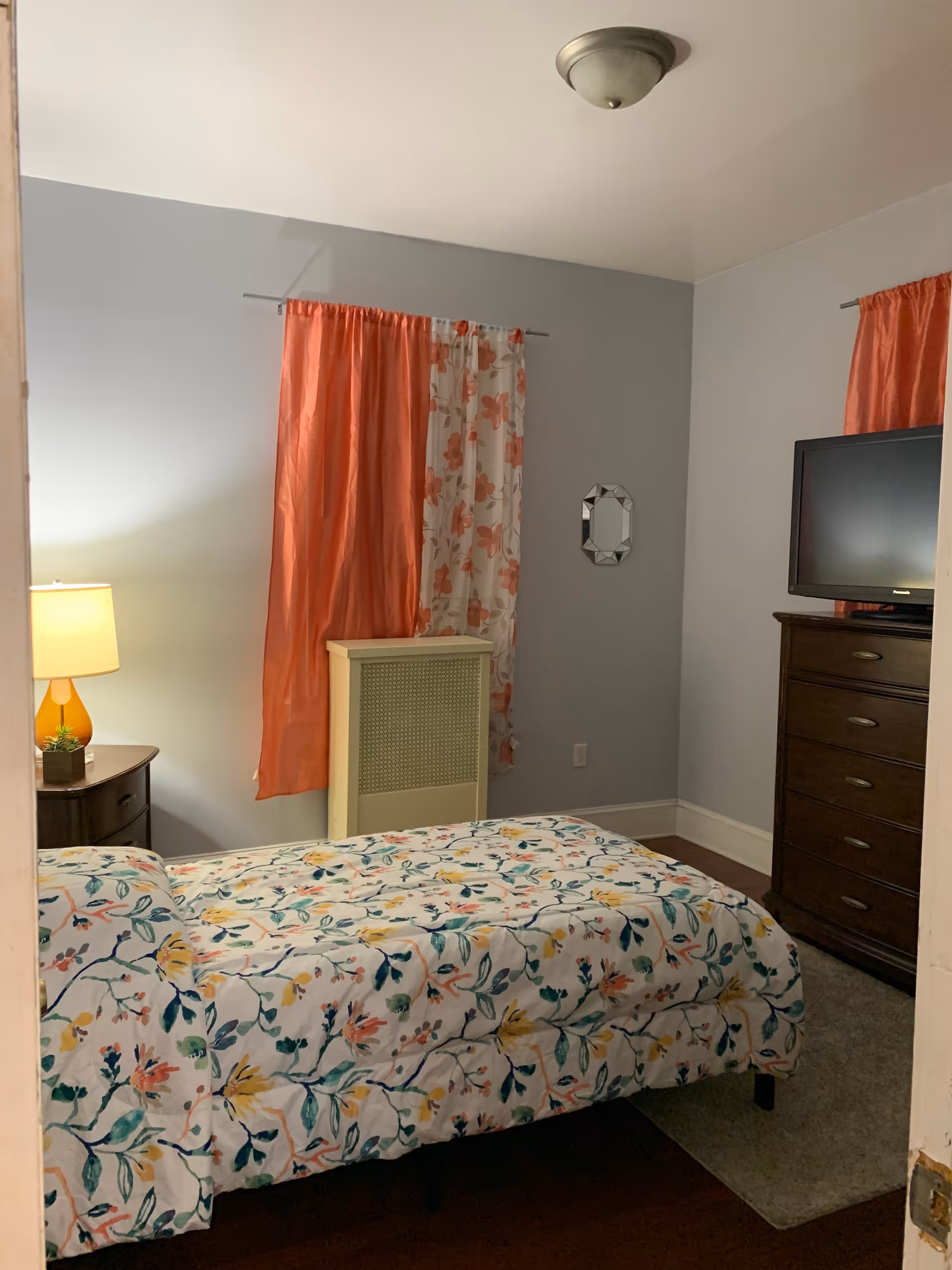 Bedroom in a senior living facility with a patterned bed, dresser, TV, and coral curtains.