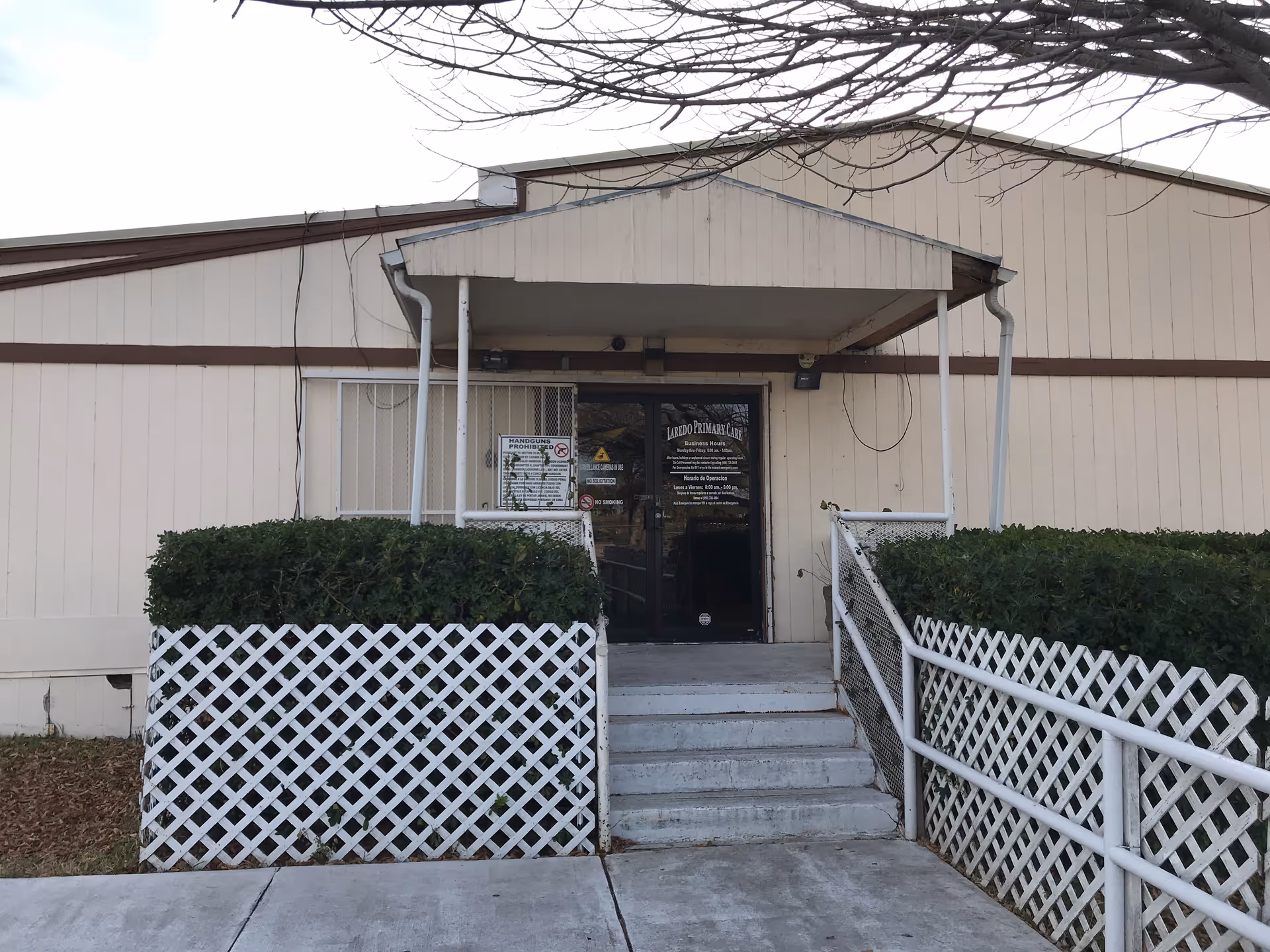 Front entrance of a beige building with a small porch, steps, and trimmed hedges.