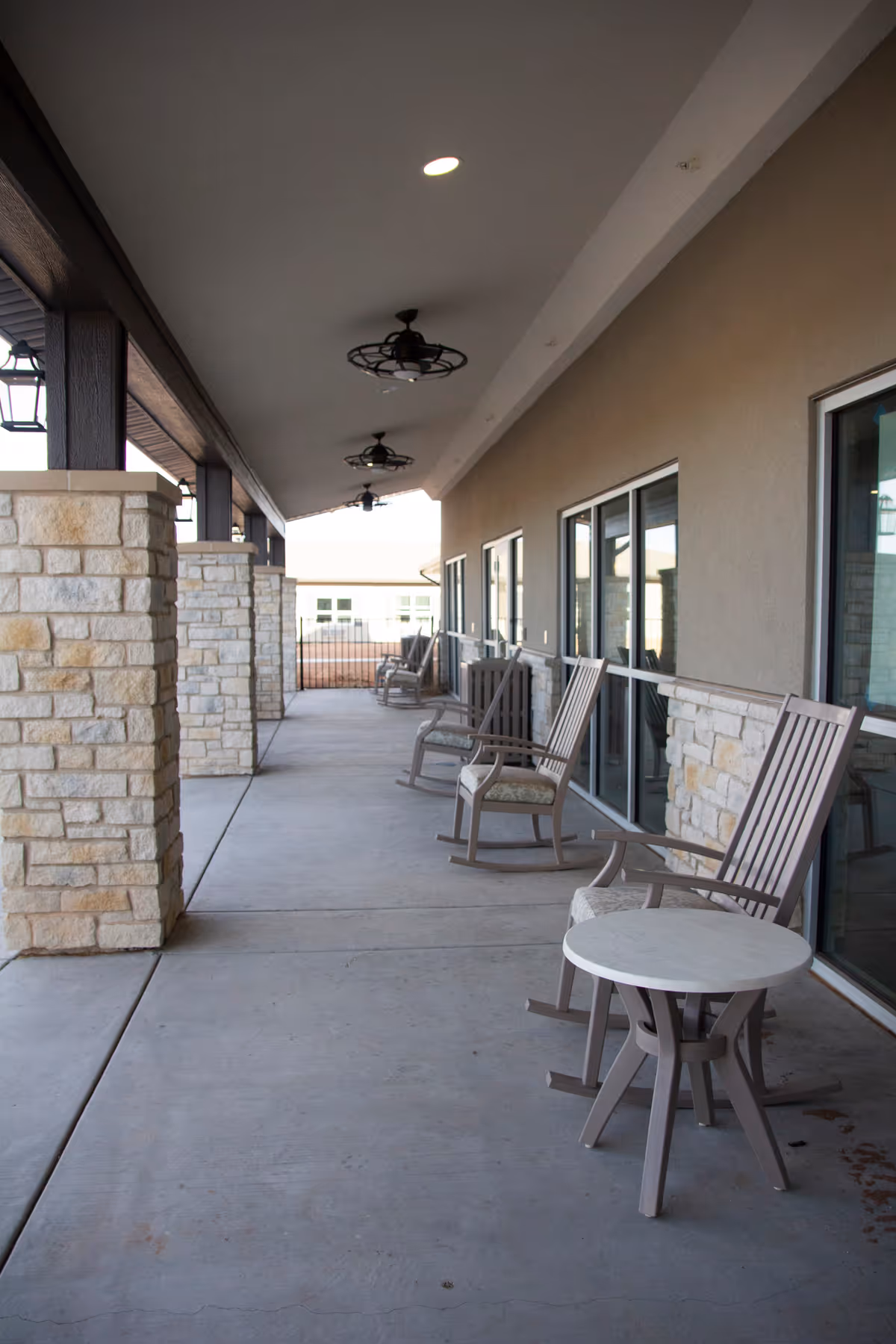 Covered outdoor patio area with stone pillars, ceiling fans, and several wooden rocking chairs with cushions arranged along the wall next to large windows. A small round table is placed near the front rocking chair.