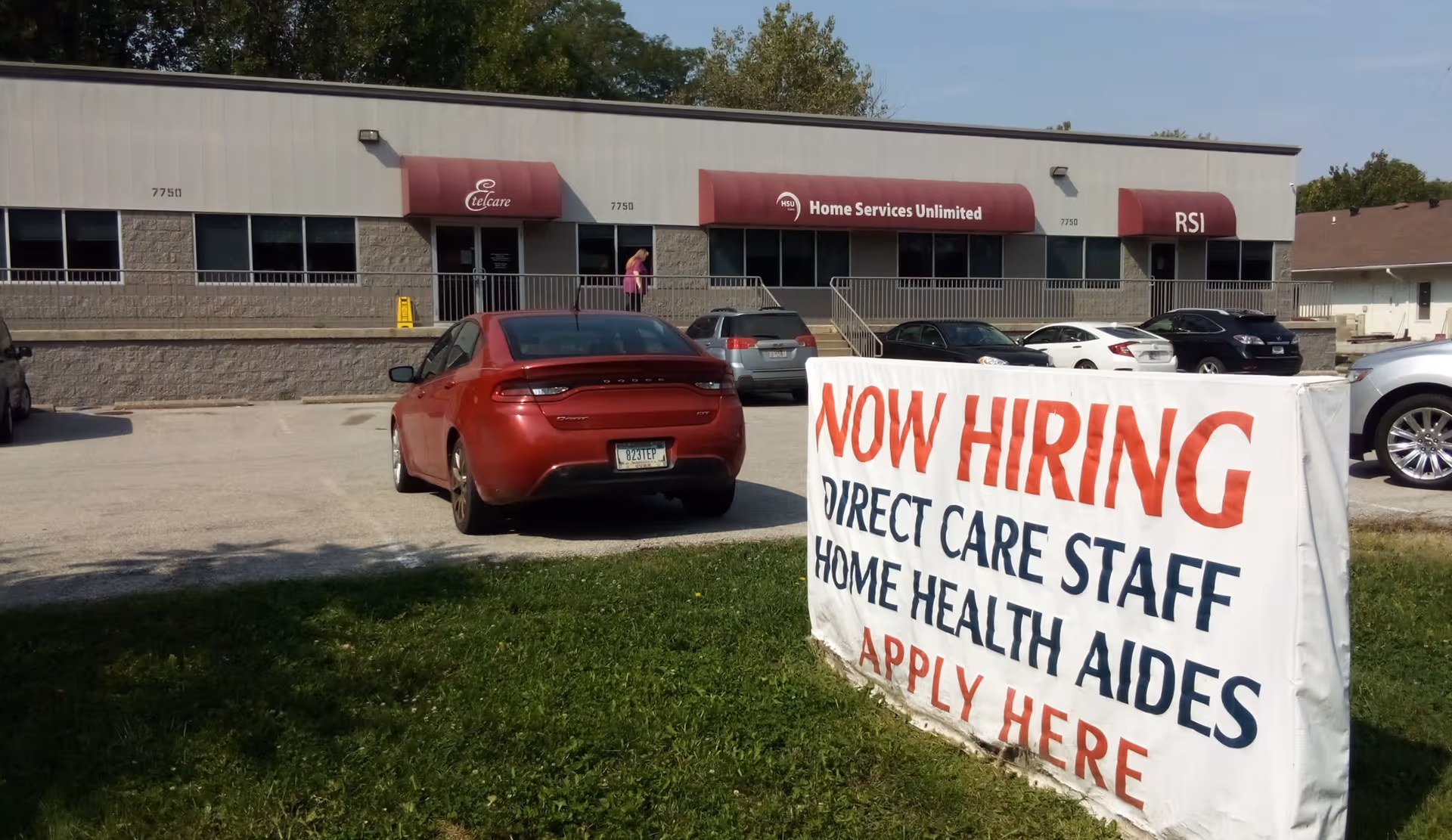 Exterior view of a care facility with a large now-hiring sign and parked cars.