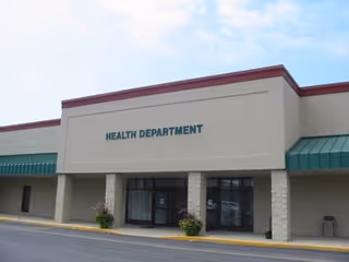 Exterior of a beige health department building with green awnings and glass entrance.