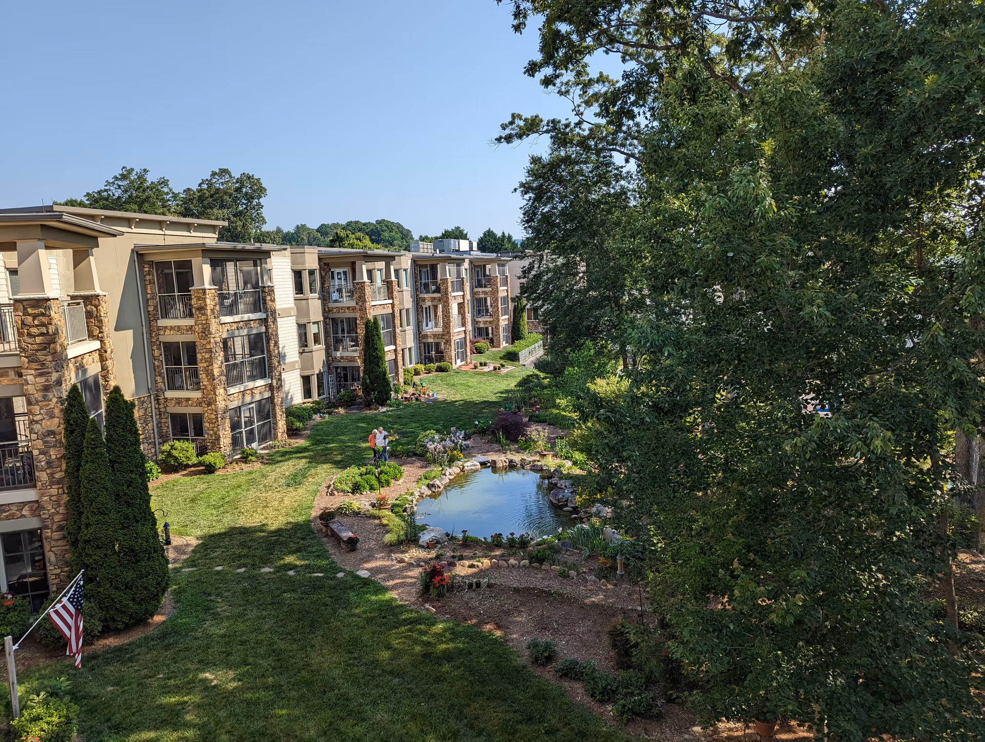 A multi-story senior living building with stone accents surrounds a landscaped courtyard and pond.