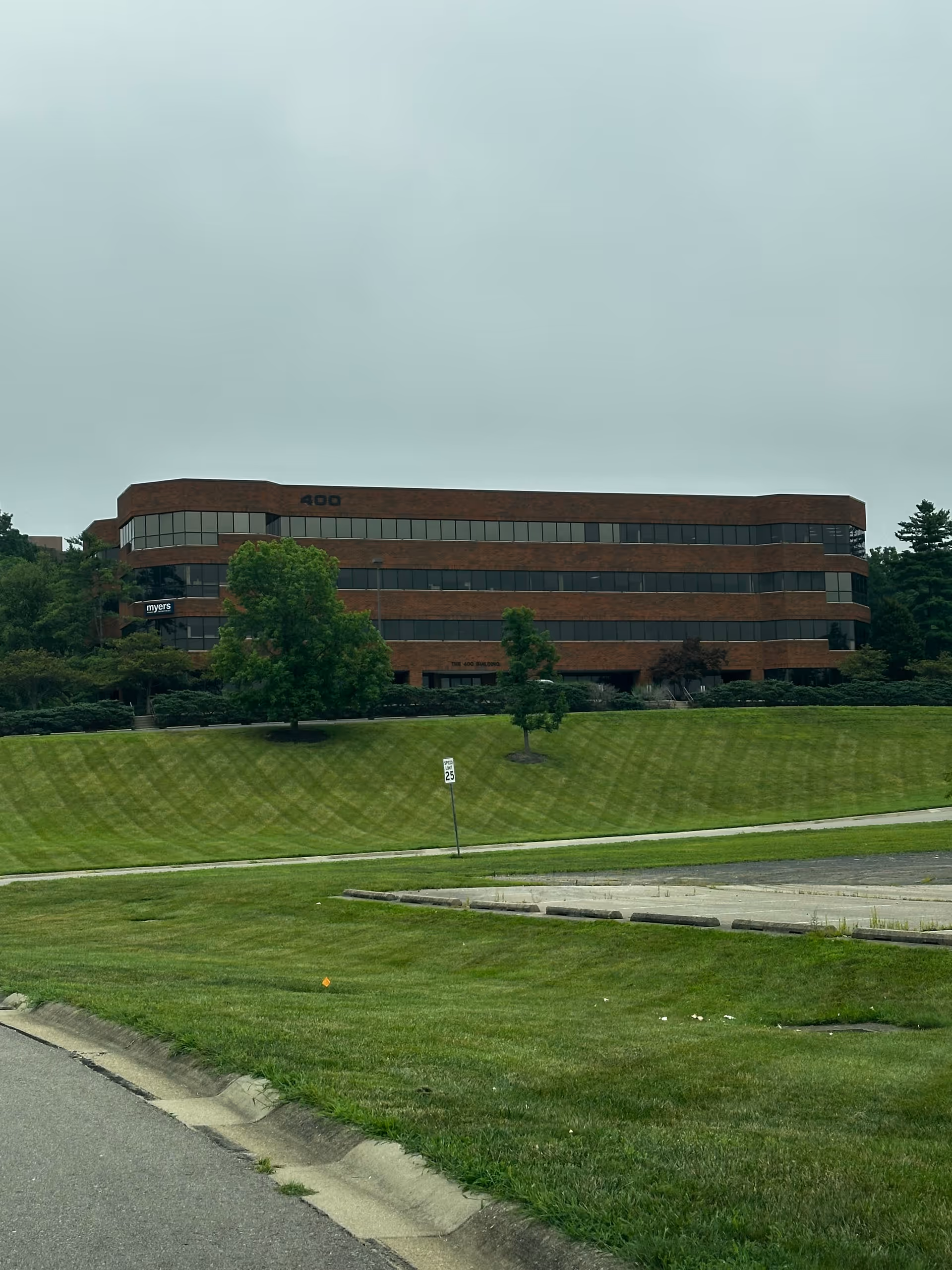 A brick multi-story office building sits atop grassy hill, with trees and a road in foreground.