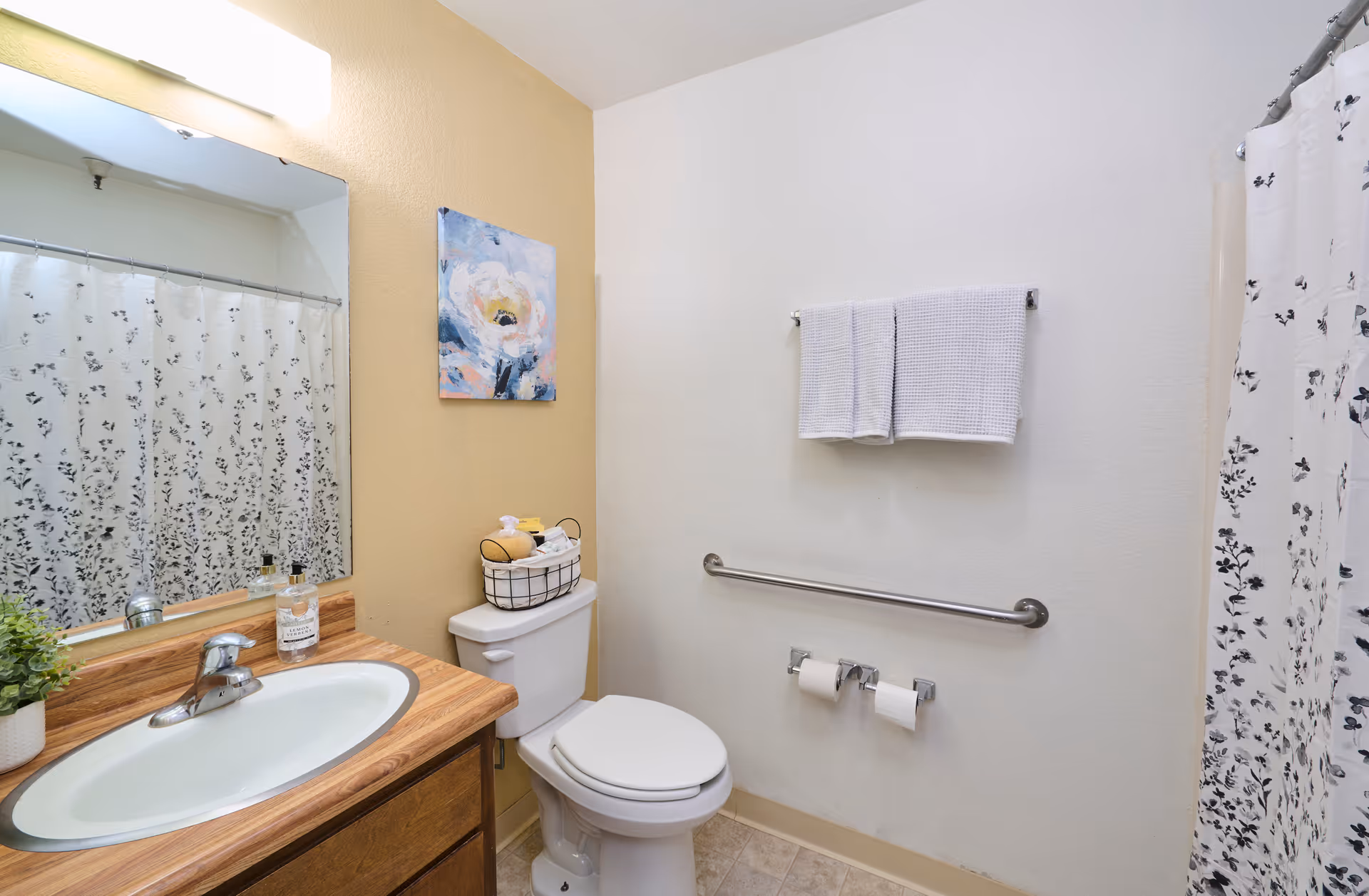 Small bathroom with a sink and wooden vanity, toilet, wall-mounted grab bar, towel rack, and floral shower curtain.