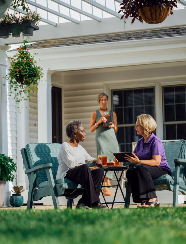 Two elderly residents chat on a sunny outdoor patio with a caregiver nearby.