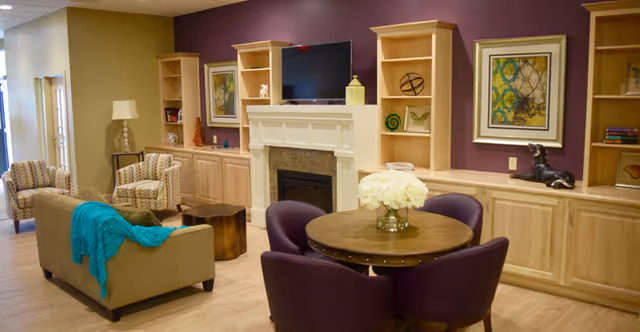Communal living room with chairs, a sofa, a round table with flowers, a fireplace under a wall-mounted TV, and built-in shelving against an accent wall.
