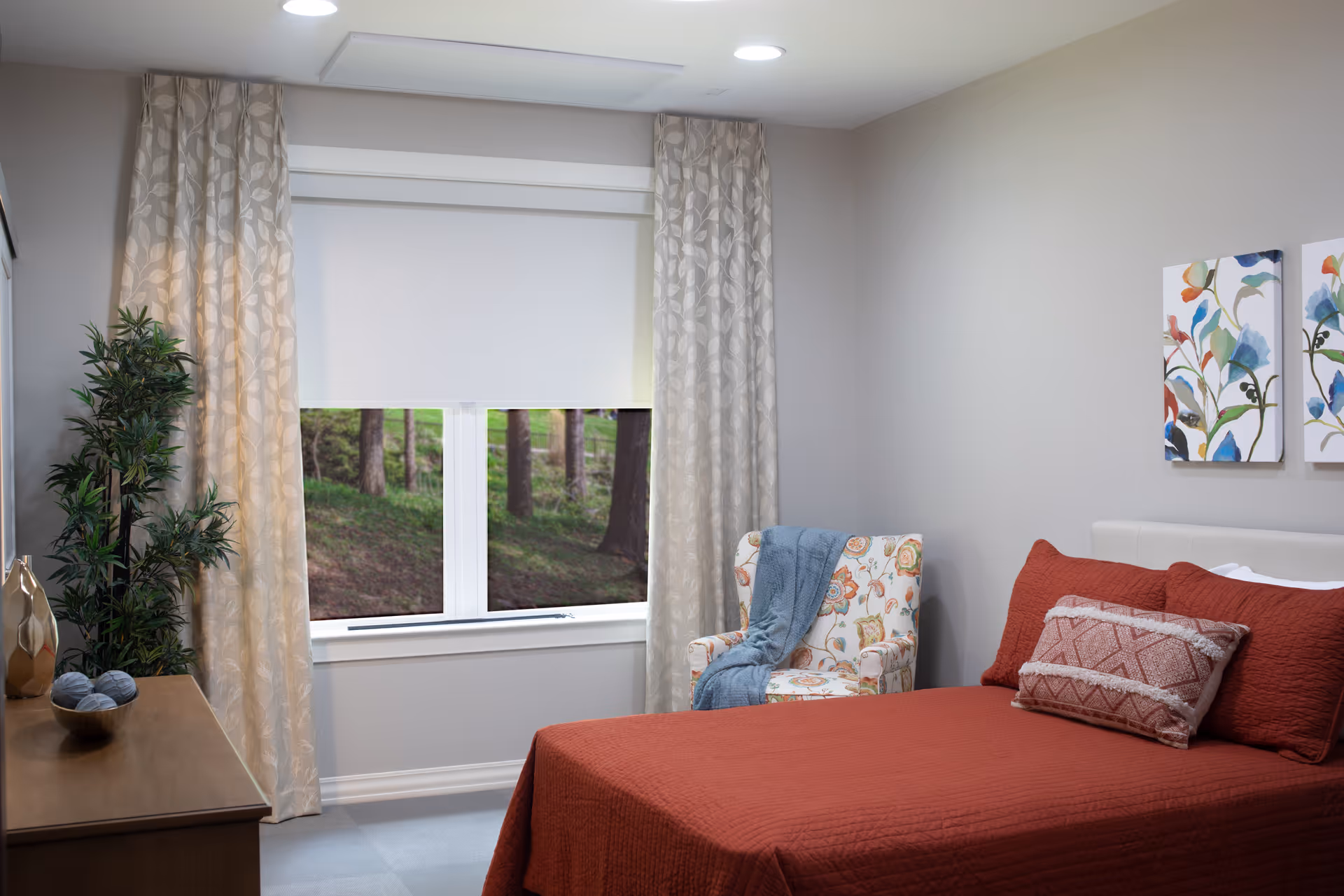 A cozy bedroom with a bed covered in a rust-colored quilt and matching pillows. There is a floral-patterned armchair with a blue throw blanket draped over it next to a window with beige curtains and a white roller shade. A wooden dresser with decorative items and a potted plant is on the left side. Two colorful floral paintings hang on the wall above the bed.