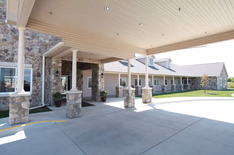 Covered entrance area of a senior living facility with stone pillars and beige siding. The driveway is concrete and there are potted plants near the entrance. The building has multiple windows and a well-maintained lawn with a small tree.
