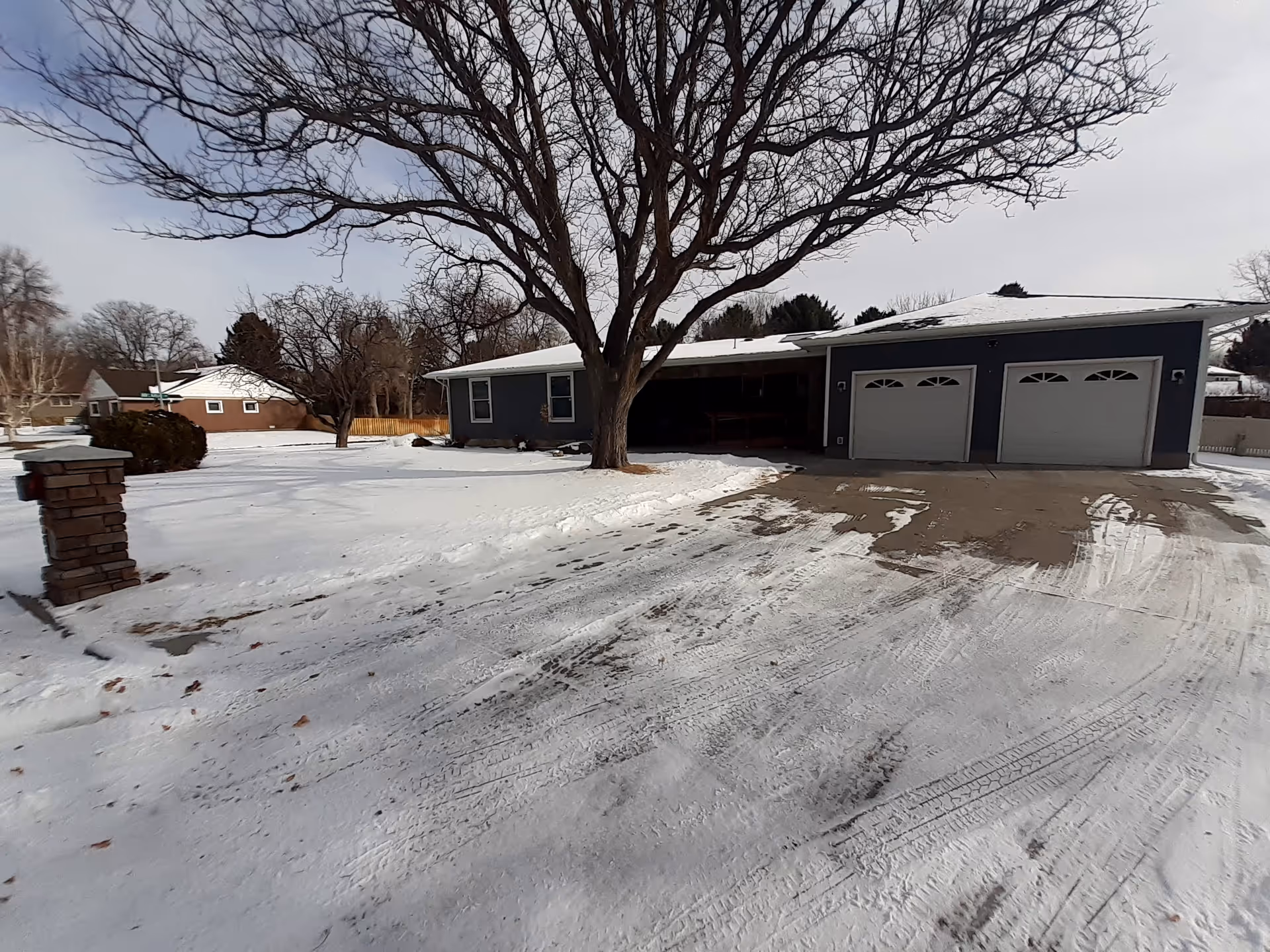 Snow covers the front of a single-story home with a two-car garage and bare trees.