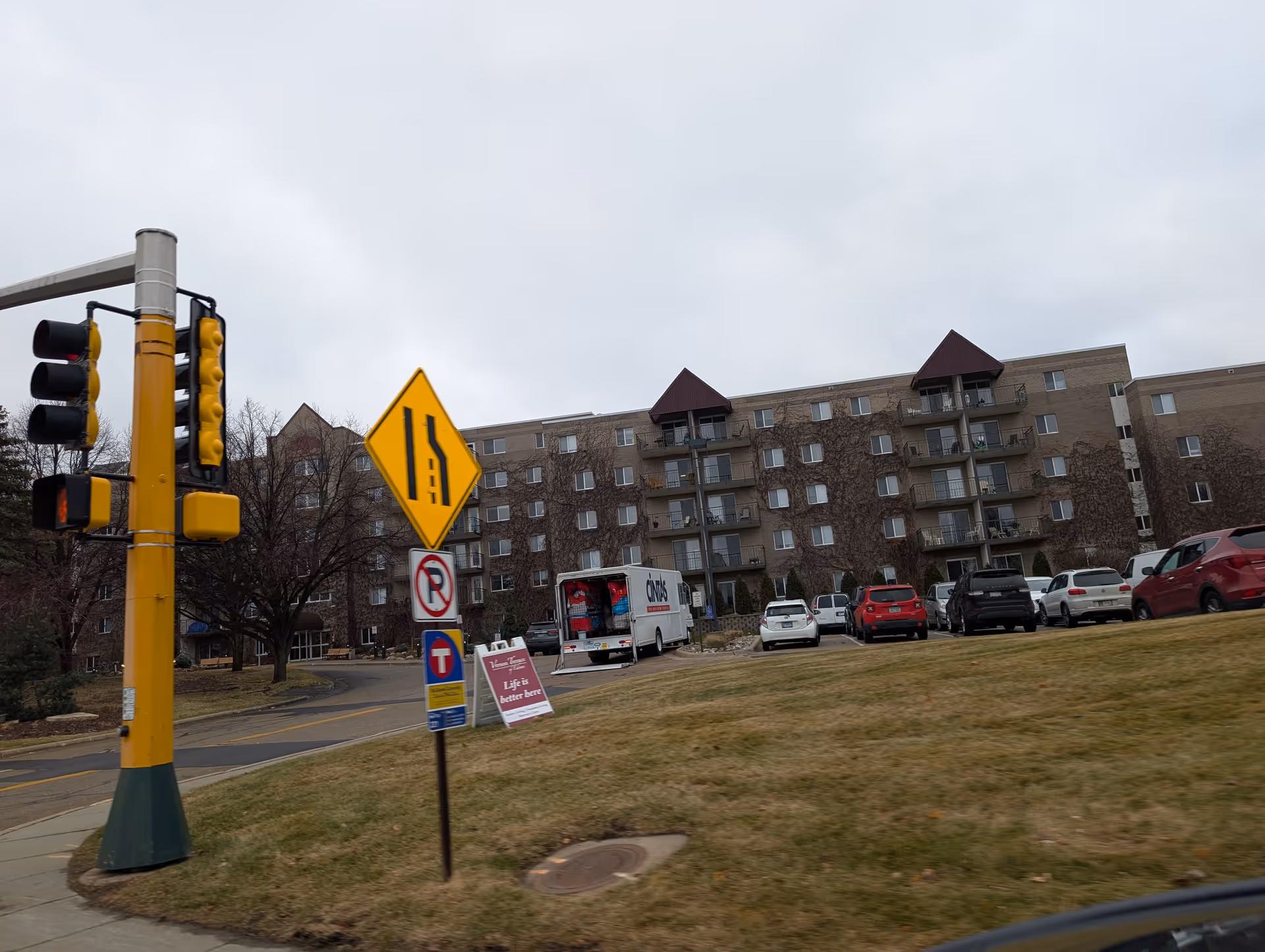 Exterior view of a multi-story senior living building with parked cars, a traffic signal, and road signs in the foreground.