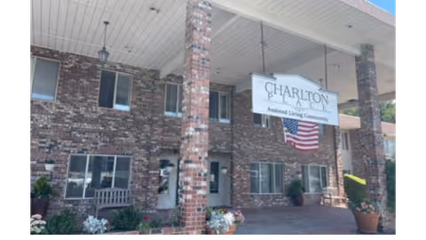 Entrance of Charlton Place Assisted Living Community showing a brick building with a covered driveway supported by brick columns. There is a large sign hanging from the ceiling with the facility name and an American flag beneath it. Potted plants and benches are placed near the entrance.