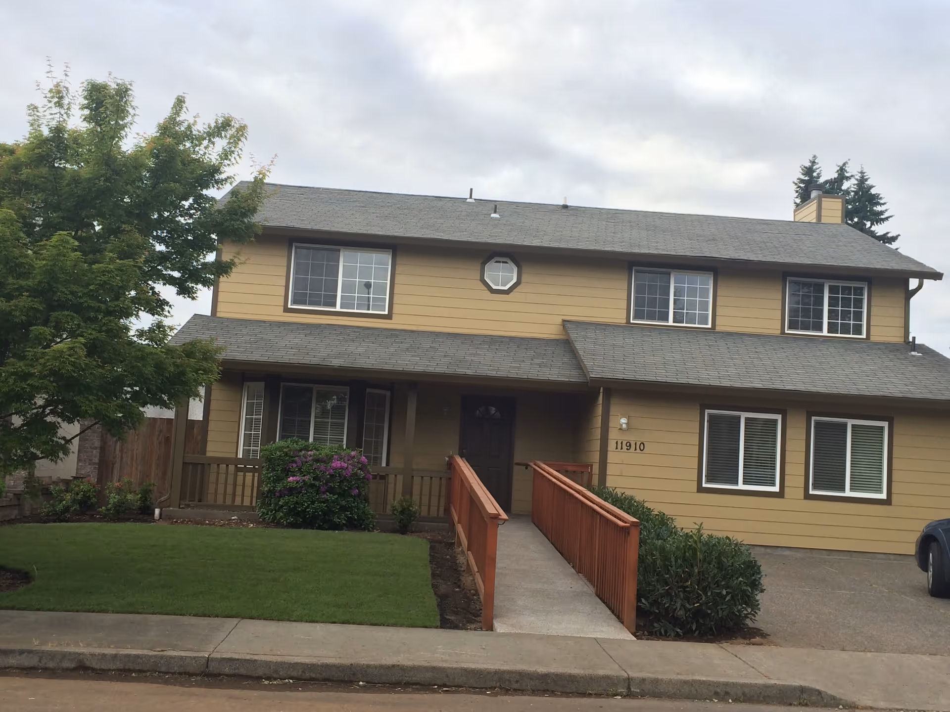 Front view of a two-story tan house with a ramped entry, porch, lawn and a tree.